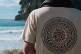 Person wearing a beige t-shirt with a circular design featuring a lion, standing on a beach. sulis
