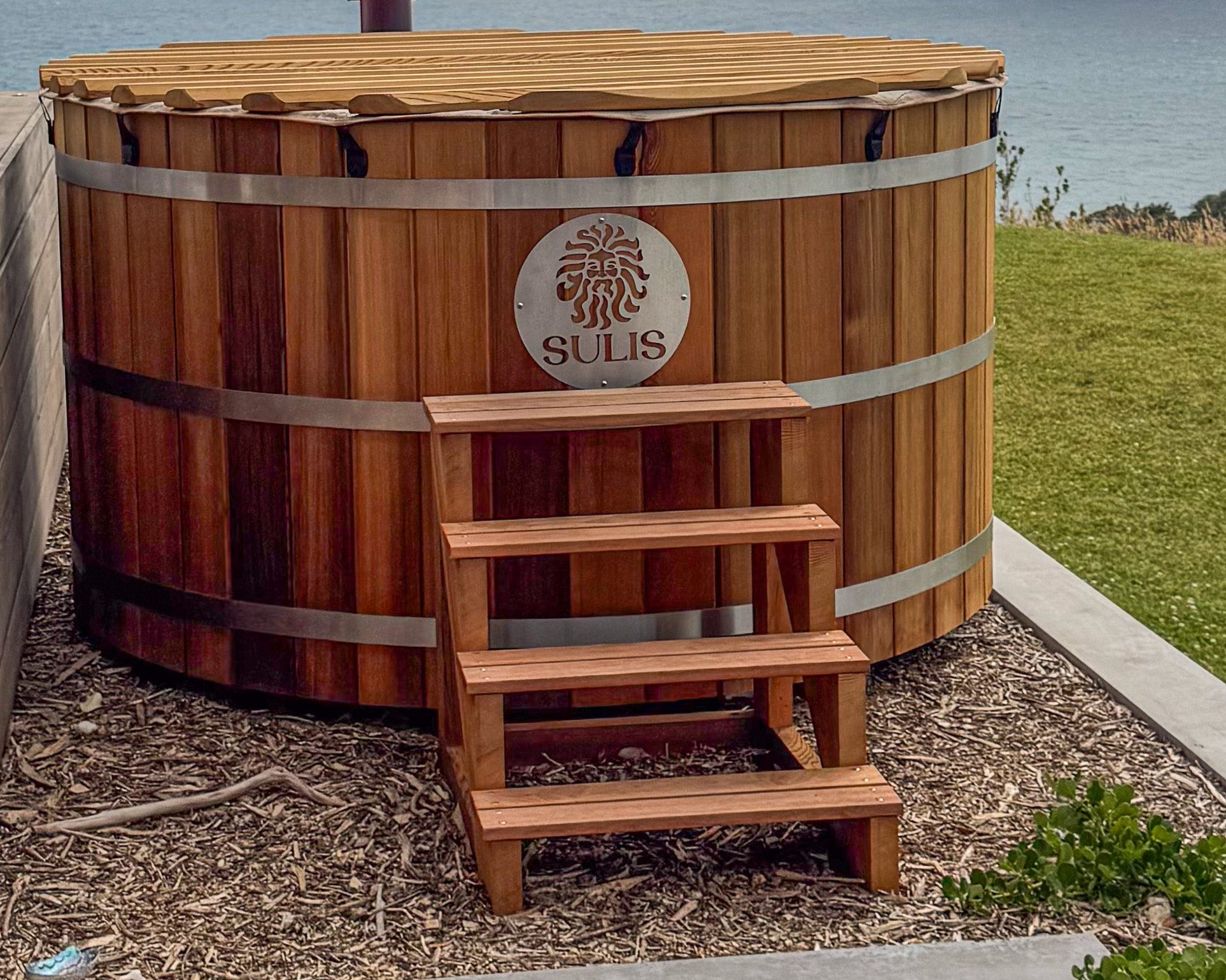 A large, outdoor hot tub made of wood, with a logo on the side, and a set of wooden steps leading up to it, set in a garden environment. Sulis Hot Tubs