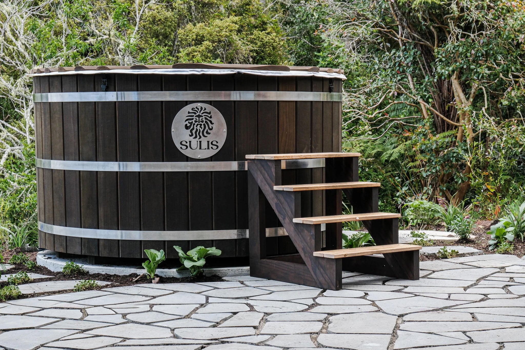 Wooden hot tub with steps in a garden setting, featuring the Sulis logo. Sulis hot Tub