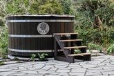Wooden hot tub with steps in a garden setting, featuring the Sulis logo. Sulis hot Tub