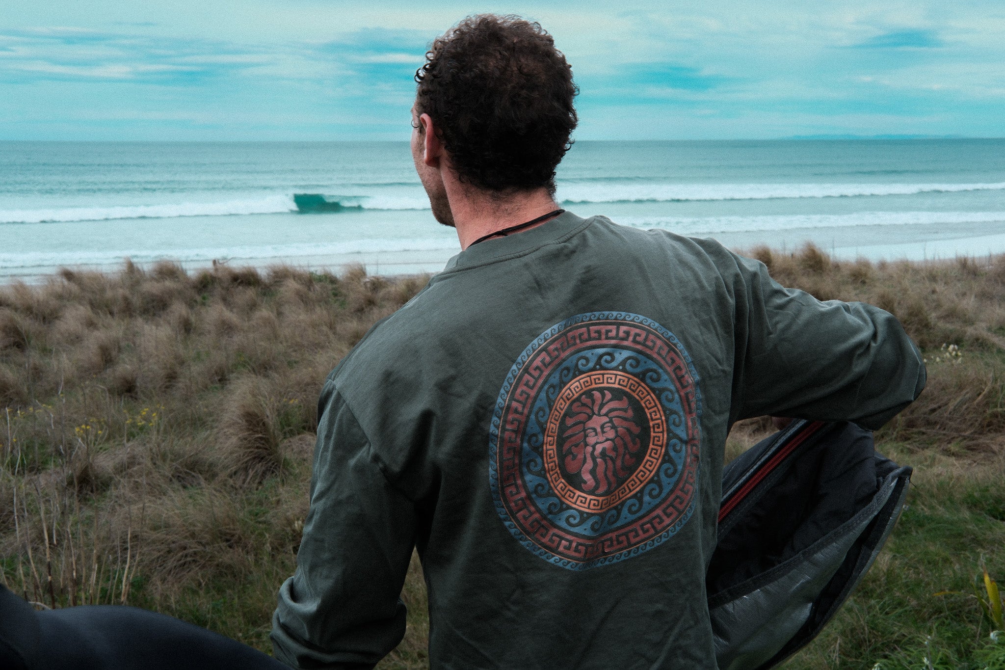 A person viewed from behind wearing a long-sleeved green t-shirt with a circular emblem design, standing in an outdoor setting with grass and a body of water in the background.