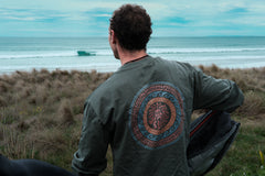 A person viewed from behind wearing a long-sleeved green t-shirt with a circular emblem design, standing in an outdoor setting with grass and a body of water in the background.