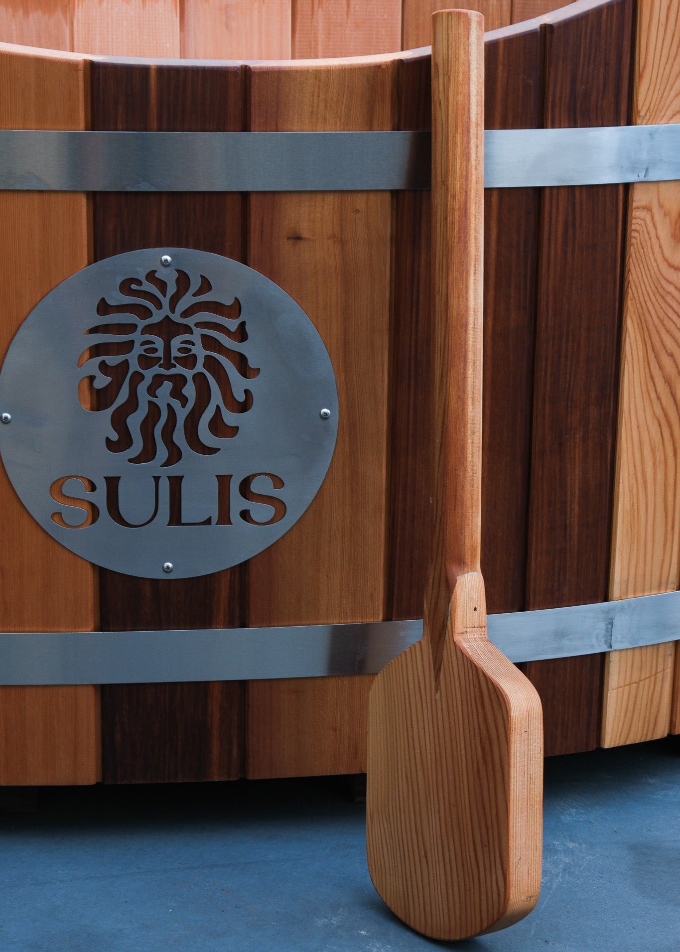 A wooden stirring paddle placed against a wooden hot tub with a metal band and the brand 'SULIS' inscribed on it. Sulis Hot Tubs