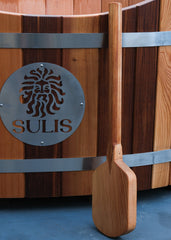 A wooden stirring paddle placed against a wooden hot tub with a metal band and the brand 'SULIS' inscribed on it. Sulis Hot Tubs