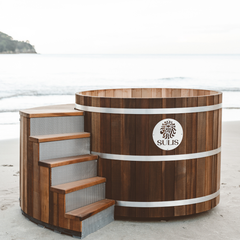 Wooden hot tub with steps on a sandy beach, featuring the Sulis logo.