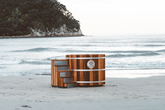 Wooden barrel-shaped structure Sulis Hot Tub on a beach with ocean and trees in the background