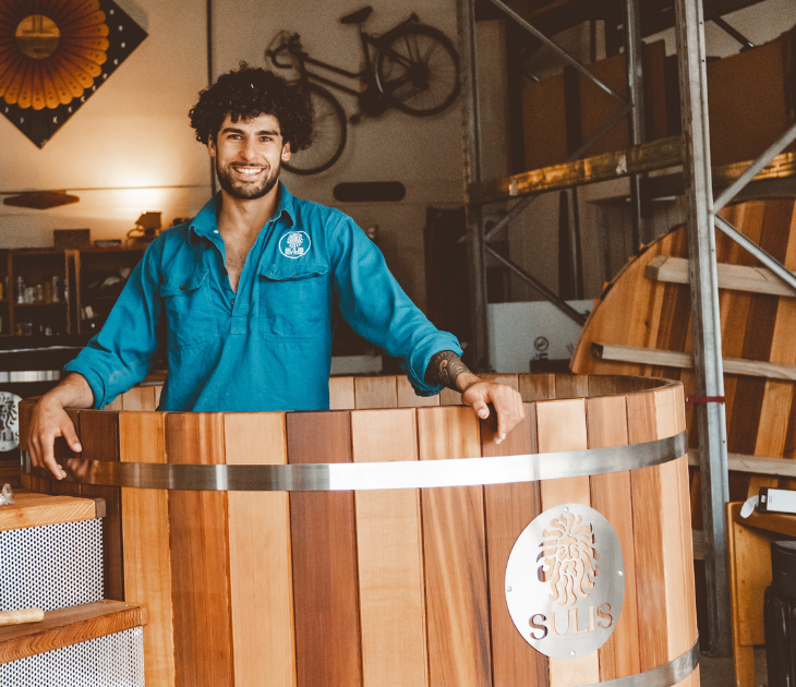 Man standing in a wooden hot tub with a logo in a rustic setting, sulis hot tubs
